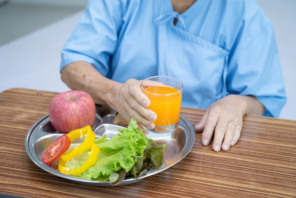 Patient in a hospital gown holding a glass of juice with a tray of fresh fruit and vegetables, a meal idea for colonoscopy recovery