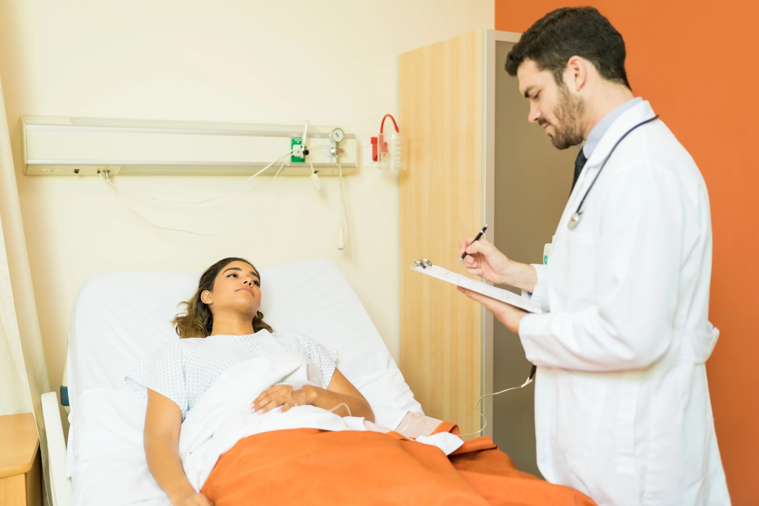 A doctor takes notes while talking with a patient in a hospital bed, supporting patient safety.